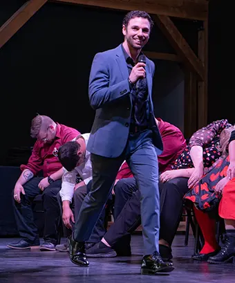 PerformancePhoto3 - Carroll News | Carroll Publications | Carroll County, MD Male singer in a blue suit performs on stage with a microphone, while a row of people seated behind him bend forward in colorful outfits.