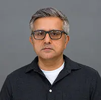 Portrait of a middle-aged man with gray hair and black-framed glasses, wearing a dark shirt against a gray background.