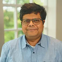 Portrait of a man wearing glasses and a light blue button-up shirt, smiling at the camera in a bright setting