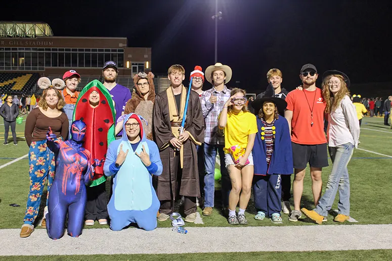 Group of friends in Halloween costumes posing on a football field at night with stadium lights in the background.