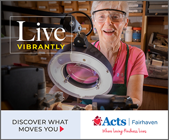 Woman in a workshop wearing safety glasses, inspecting a device with a circular lamp; features the slogan 'Live Vibrantly' and a bottom banner reading 'Discover What Moves You' with the Acts Fairhaven logo.