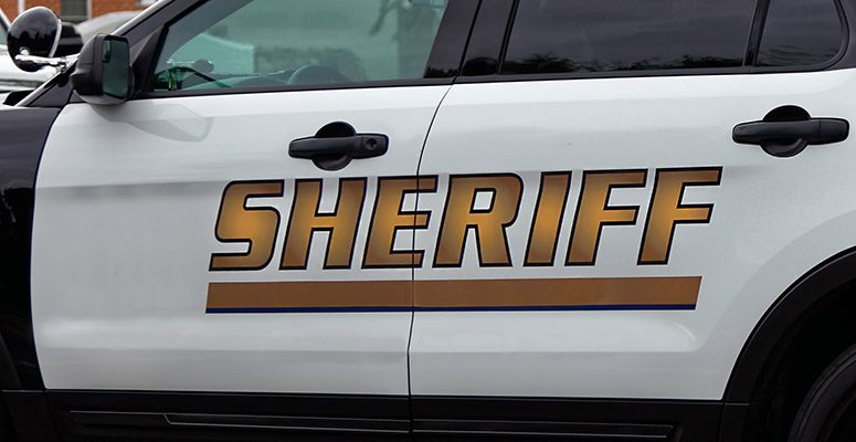 Side of a white sheriff patrol SUV with large gold SHERIFF lettering and blue stripe on the door surface of the vehicle, front and rear doors visible.