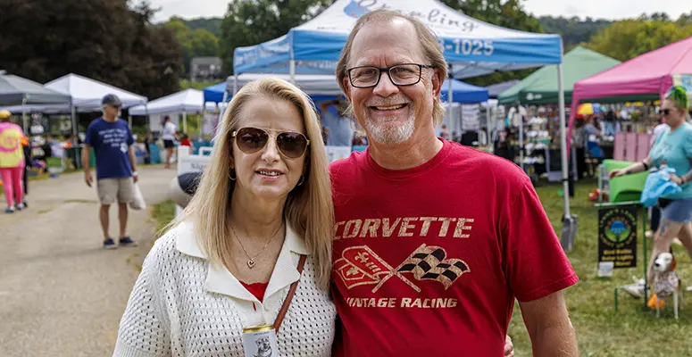 Smiling couple posing at an outdoor festival with colorful vendor tents in the background.