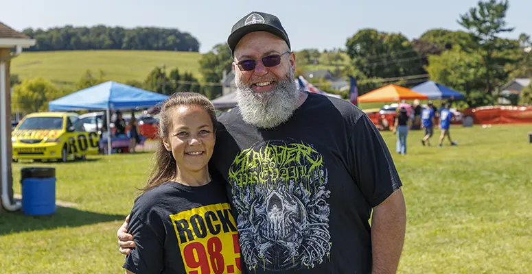 Smiling couple posing outdoors at a festival, wearing Rock 98.9 shirts, with tents and a yellow van in the background.