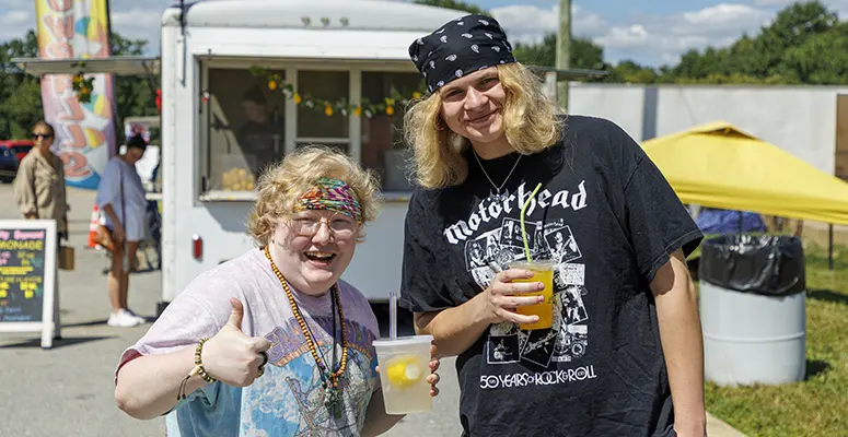 Two smiling people holding cups at an outdoor festival, giving a thumbs-up to the camera.