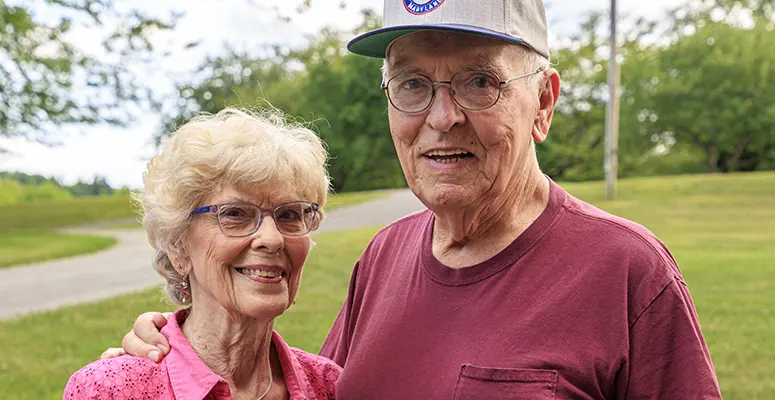 Smiling elderly couple standing close together in a park, arms around each other on a sunny day.
