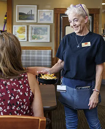 Smiling server in a navy shirt hands a plate of fries to a seated woman in a cozy dining area.