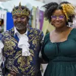 Two people pose at a costume market: a man in a royal blue and gold outfit with crown and scepter, and a woman in a green dress with glasses.