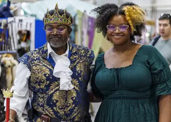 Two people pose at a costume market: a man in a royal blue and gold outfit with crown and scepter, and a woman in a green dress with glasses.