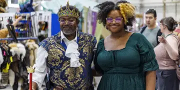 Two people pose at a costume market: a man in a royal blue and gold outfit with crown and scepter, and a woman in a green dress with glasses.