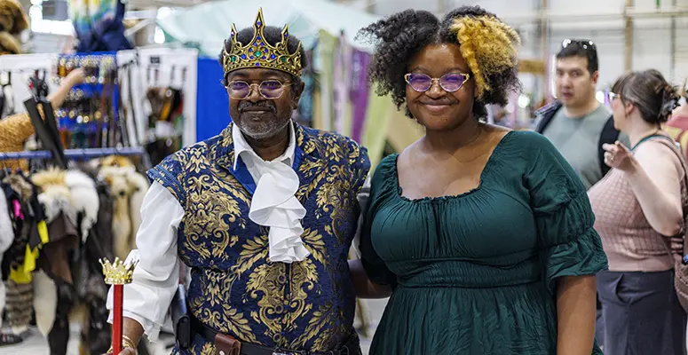 Two people pose at a costume market: a man in a royal blue and gold outfit with crown and scepter, and a woman in a green dress with glasses.