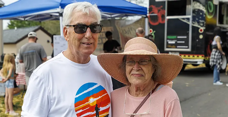 Senior man and woman wearing sunglasses pose together at an outdoor festival with food trucks in the background and tents overhead.
