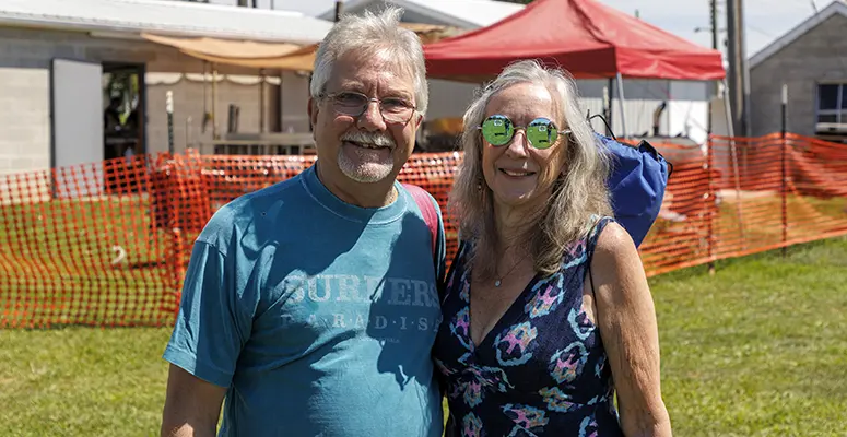 Smiling older man and woman stand together at an outdoor event with orange safety fencing and tents behind them.