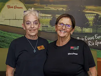 Two smiling female staff members in black shirts with name badges pose together in front of a farm-m themed mural or backdrop.