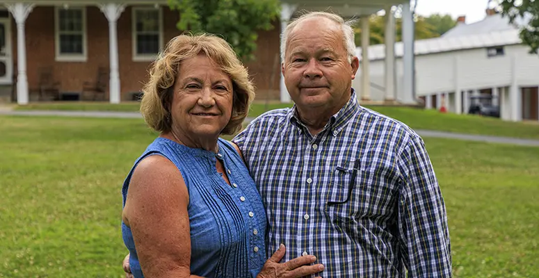 Older couple standing on a grassy lawn, arm in arm, smiling at the camera with buildings in the background.