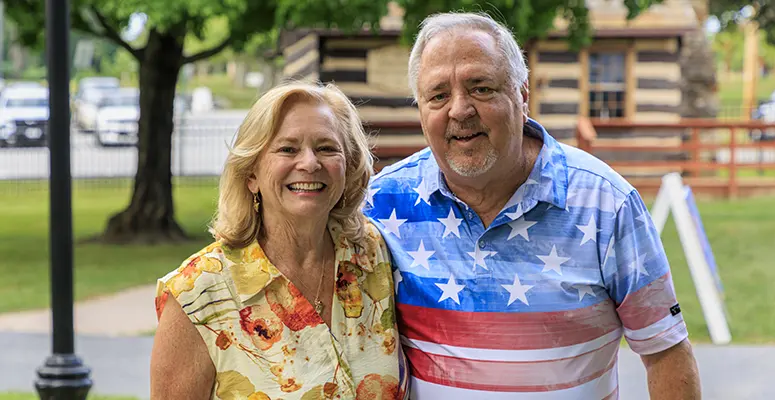 Smiling older couple standing side by side in a park, wearing colorful casual shirts and looking at the camera.