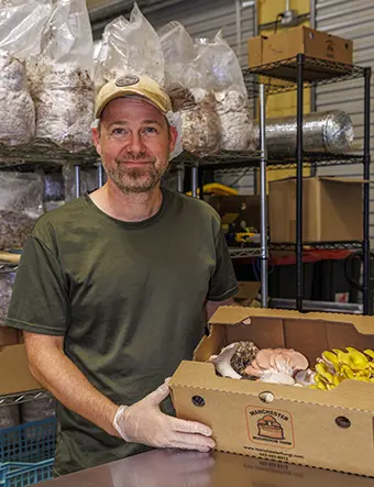 Smiling man in a cap and gloves packs a cardboard box of mushrooms in a warehouse.