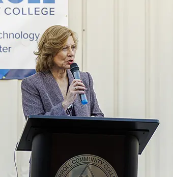 Woman in a gray blazer speaks into a microphone at a podium with a college seal.