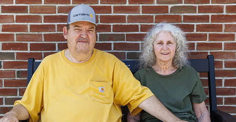 Two older adults sitting on a bench against a brick wall; man in a yellow shirt and cap, woman in a green shirt.