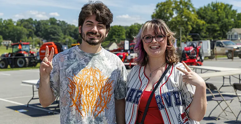 Two smiling friends stand outdoors, both making peace signs at the camera during a sunny event.