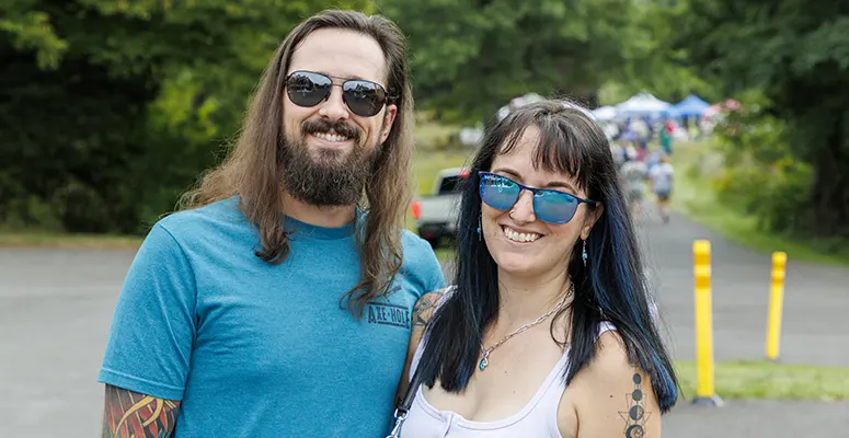 Smiling couple wearing sunglasses outdoors at a park event, with tents in the background.