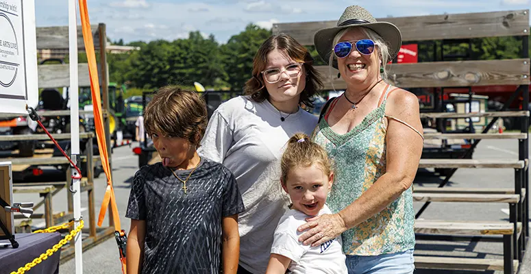 A smiling family of four posing at an outdoor fair or market, with a woman in a sunhat and sunglasses beside two girls and a boy in front of wooden bleachers.