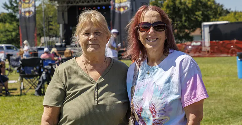 Two women posing together at an outdoor event on a sunny day, smiling near a stage in the background.