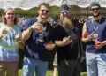 Group of four men standing under a white tent at an outdoor festival, holding drinks and snacks and posing for the camera.