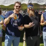 Group of four men standing under a white tent at an outdoor festival, holding drinks and snacks and posing for the camera.