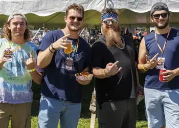 Group of four men standing under a white tent at an outdoor festival, holding drinks and snacks and posing for the camera.