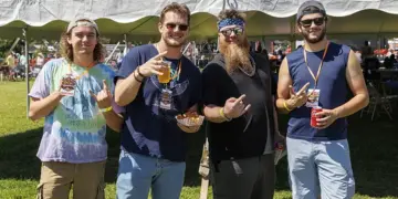 Group of four men standing under a white tent at an outdoor festival, holding drinks and snacks and posing for the camera.