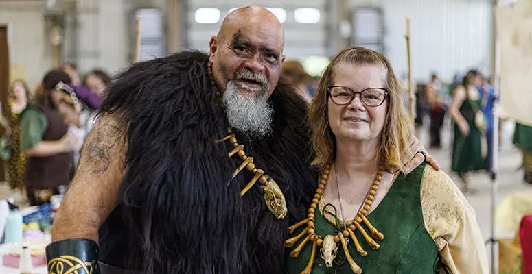 Two adults in fantasy costumes pose for a photo at a convention; man in a fur cloak with bone necklace and beard, woman in a green medieval dress.