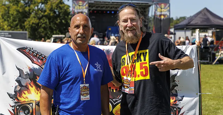 Two men posing at an outdoor festival with a stage in the background; one in a blue shirt with a lanyard, the other in a black 'Rocky 99.5' shirt.