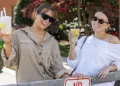 Two women wearing sunglasses lean on a wooden fence, holding lemonades and smiling outdoors at a sunny event.",