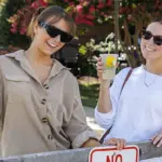 Two women wearing sunglasses lean on a wooden fence, holding lemonades and smiling outdoors at a sunny event.",
