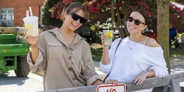Two women wearing sunglasses lean on a wooden fence, holding lemonades and smiling outdoors at a sunny event.",