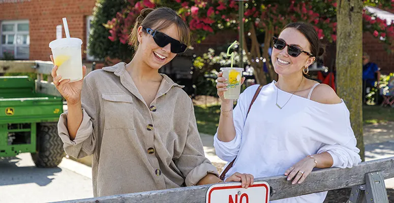 Two women wearing sunglasses lean on a wooden fence, holding lemonades and smiling outdoors at a sunny event.",