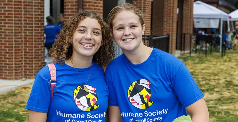 Two smiling volunteers in blue Humane Society t-shirts stand together outdoors at an event.