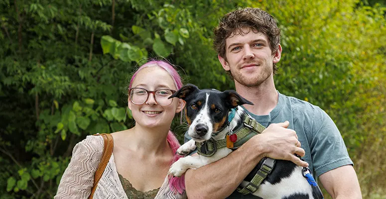 Couple outdoors with their black-and-white dog wearing a harness, all smiling at the camera.