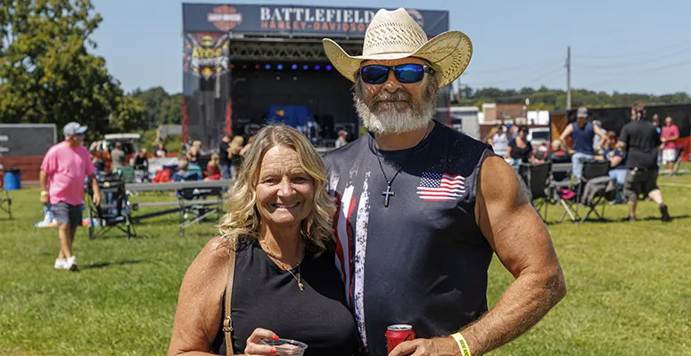Smiling couple with drinks at an outdoor festival; man in a cowboy hat and USA flag sleeveless shirt, woman in a black top, stage behind them.