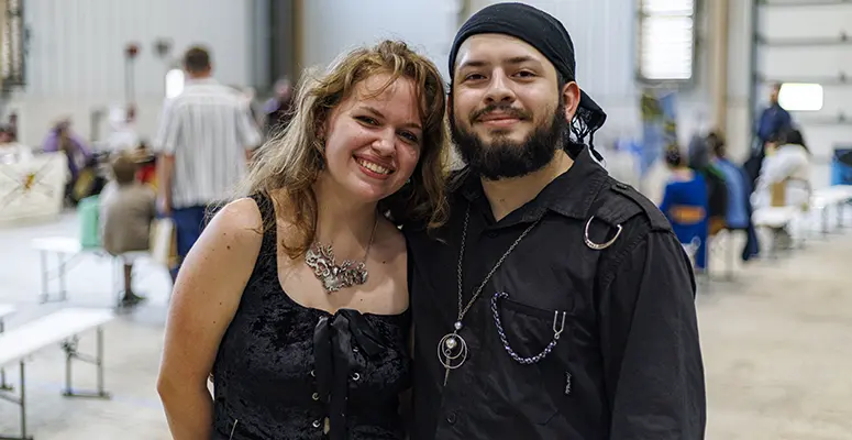 Smiling woman with wavy blonde hair and a bearded man in a black shirt posing together at an indoor event with people in the background.