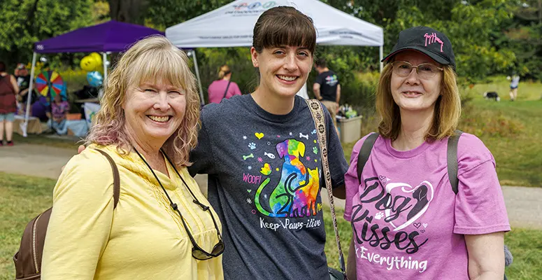 Three women posing together at an outdoor community event in a park, with tents in the background and everyone smiling.