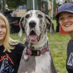 Girl in a Metallica shirt poses with a large Great Dane and a smiling woman at an outdoor festival in a park.
