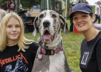 Girl in a Metallica shirt poses with a large Great Dane and a smiling woman at an outdoor festival in a park.