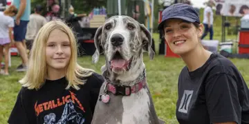Girl in a Metallica shirt poses with a large Great Dane and a smiling woman at an outdoor festival in a park.