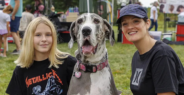 Girl in a Metallica shirt poses with a large Great Dane and a smiling woman at an outdoor festival in a park.