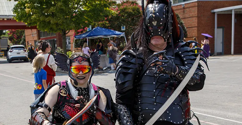 Two cosplayers in black armor wielding swords pose together at an outdoor event, one with a horned helmet and red visor.