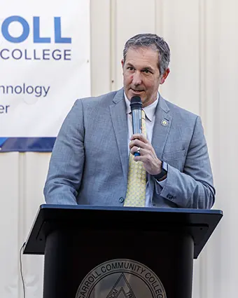 Man in a blue suit giving a speech at a podium with a microphone, standing in front of a banner that reads college