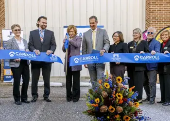 Group of ten adults in business attire participate in a ribbon-cutting ceremony for Carroll Community College, with a blue ribbon and a colorful flower arrangement at the front.