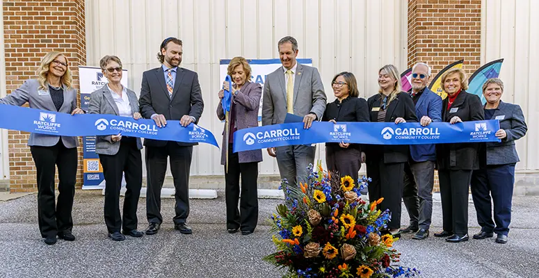 Group of ten adults in business attire participate in a ribbon-cutting ceremony for Carroll Community College, with a blue ribbon and a colorful flower arrangement at the front.